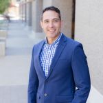 Mike Mendez standing in front of building pathway wearing blue suit jacket, checked blue and white dress shirt, and tan pants.